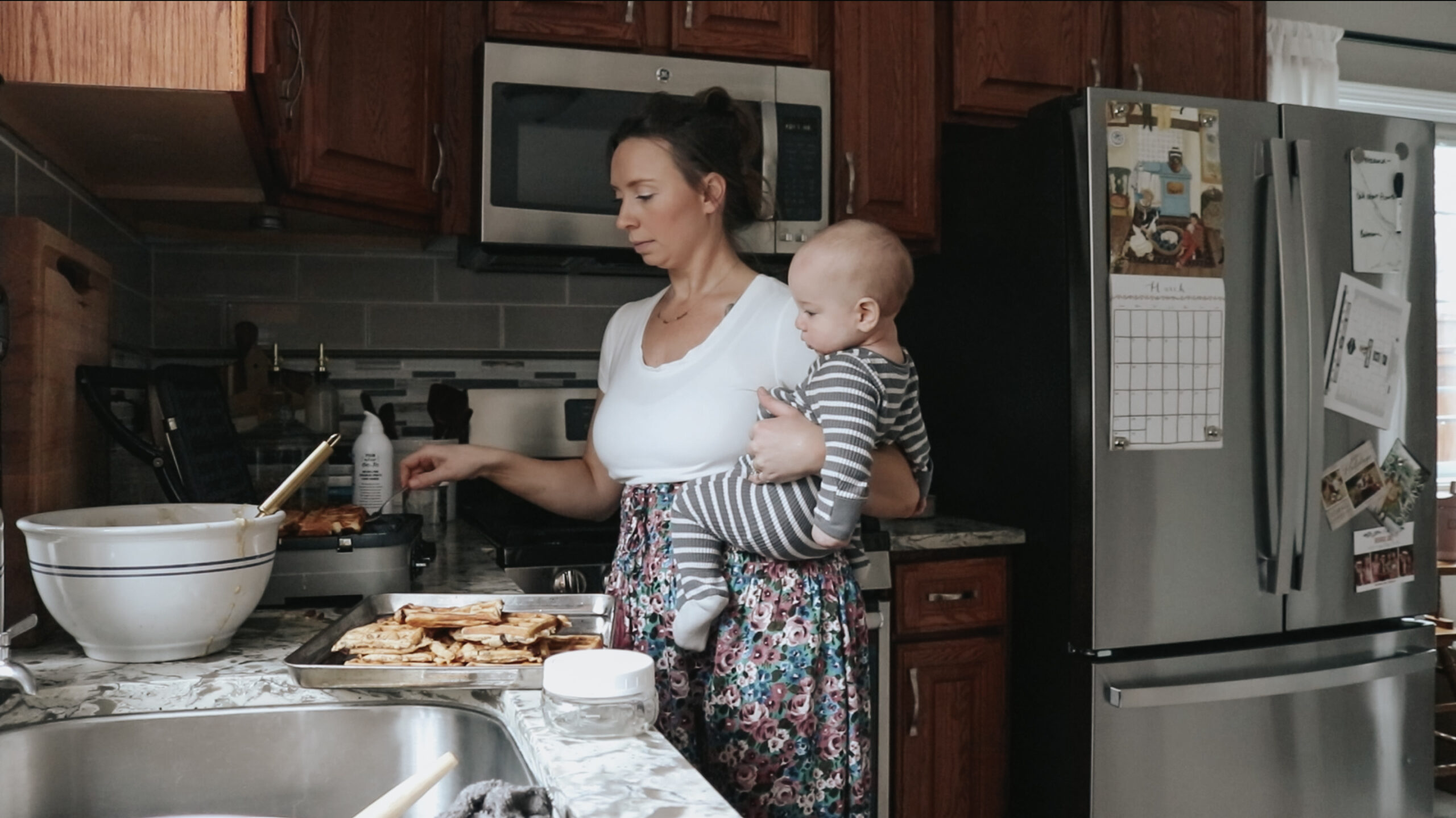 Christian mom holds her baby at the kitchen counter. She is making treats for her children while wearing a white top with a long floral skirt.